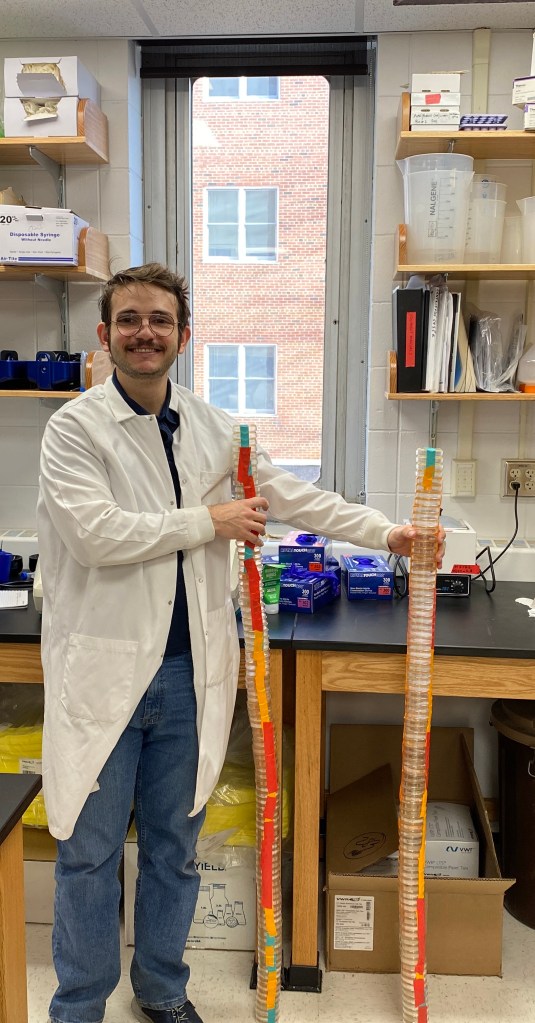 A smiling researcher in a lab coat stands in a laboratory holding two tall stacks of Petri dishes filled with various colored agar, with lab equipment visible on a counter in the background.